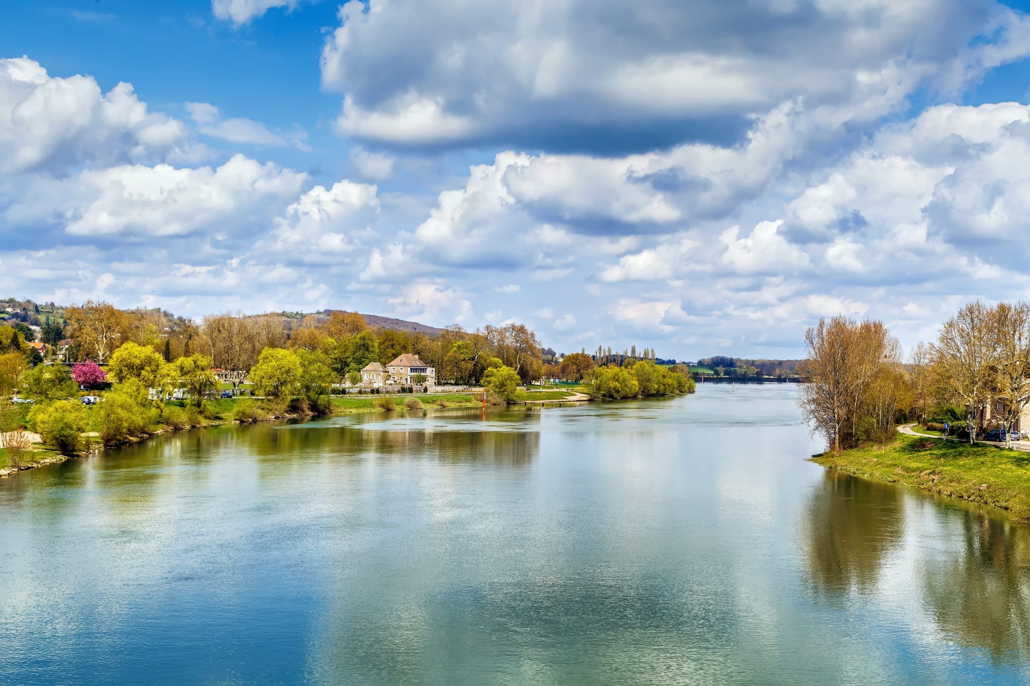 Riverside scene with calm water, trees in spring foliage, and houses lining the riverbank under a partly cloudy blue sky.