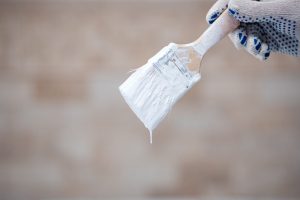 Gloved hand holding a paintbrush with white paint dripping from the bristles against a neutral blurred background