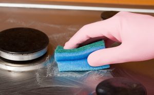 Pink-gloved hand wiping a stainless steel stovetop with a blue sponge on a reflective surface.