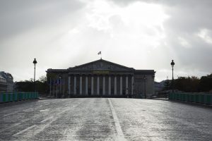 View along a cobblestone street toward the Palais Bourbon, the French National Assembly building with a flag atop and classical columns, flanked by lampposts under a cloudy sky.