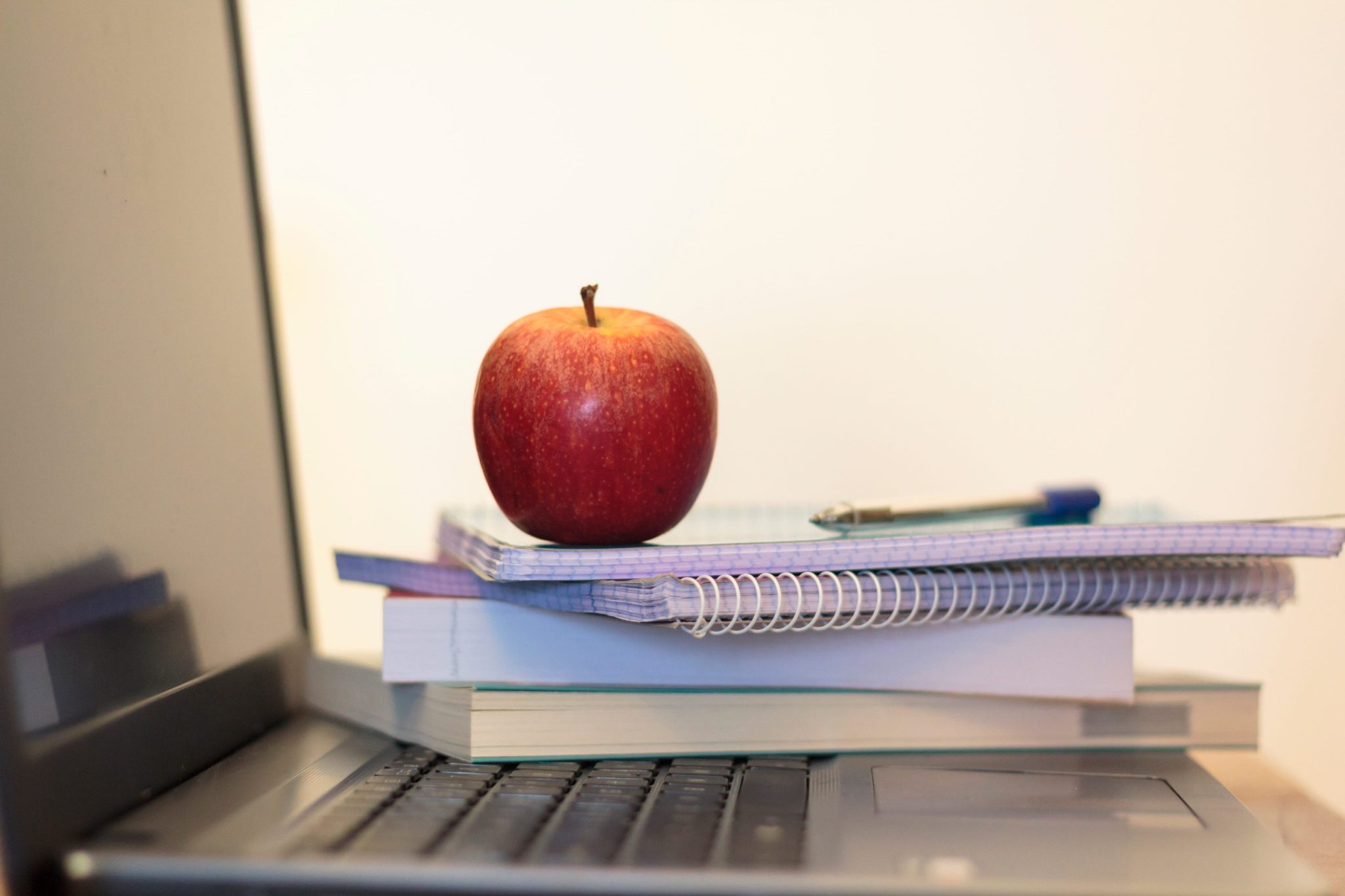 Red apple perched on top of a stack of spiral notebooks resting on a laptop keyboard, in a desk setup