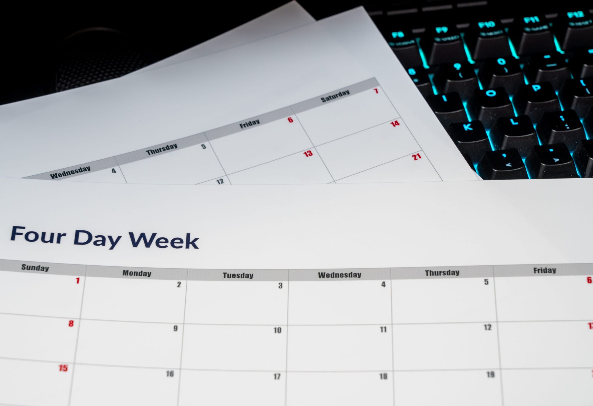 Close-up of a printed monthly calendar page with a four-day-week header, calendar grid and red dates, with a backlit keyboard in the background