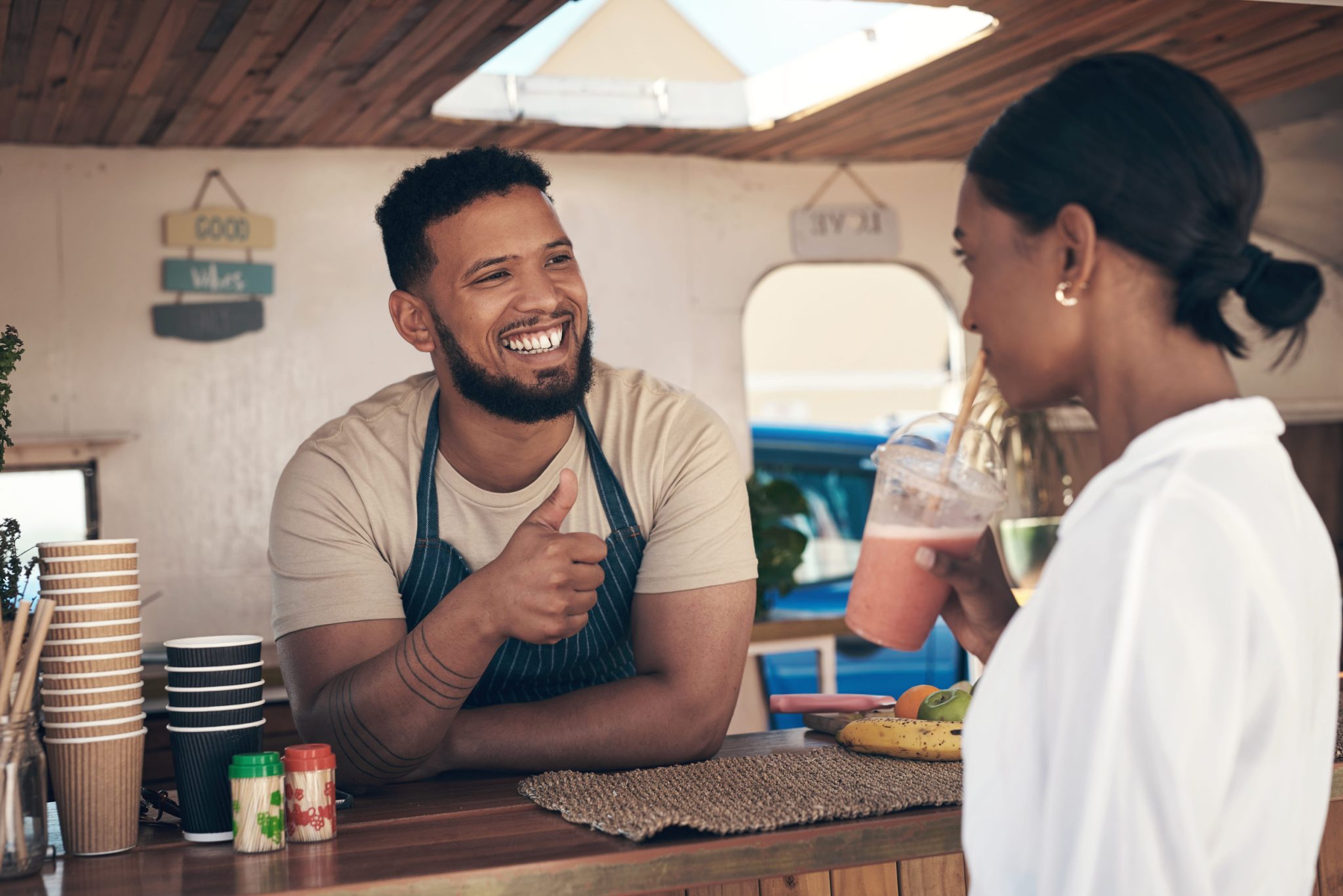 Smiling man in an apron gives a thumbs-up while talking to a woman at a cafe counter with stacked cups and fruit nearby.