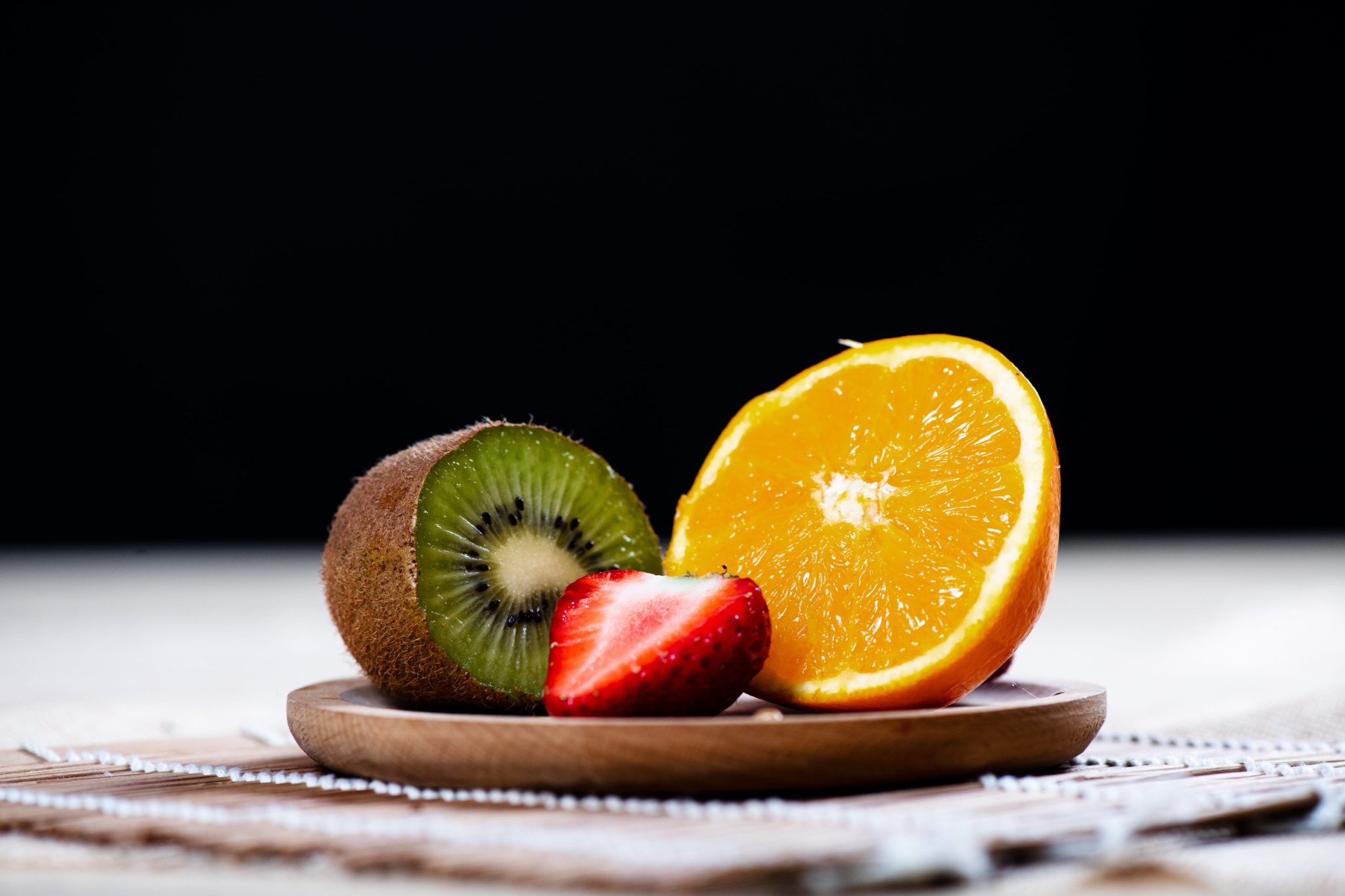 Close-up of kiwi half, orange half, and strawberry on a wooden plate against a dark background, showcasing fresh fruit.