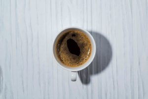 Top-down view of a white cup of black coffee on a pale blue wooden surface with visible grain patterns and a shadow to the right.