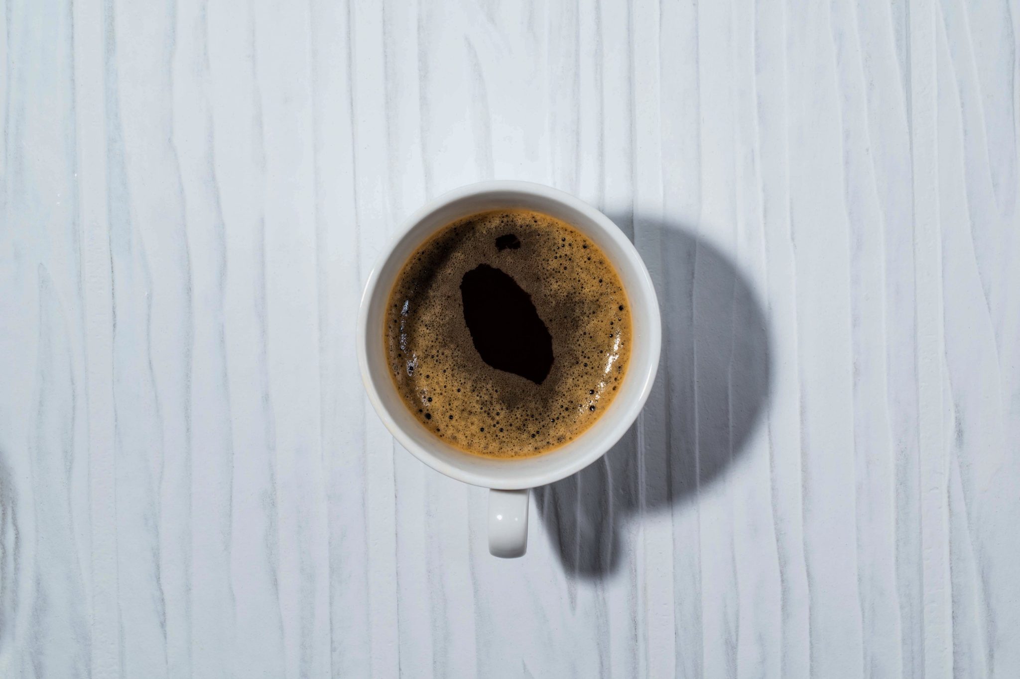 Top-down view of a white cup of black coffee on a pale blue wooden surface with visible grain patterns and a shadow to the right.