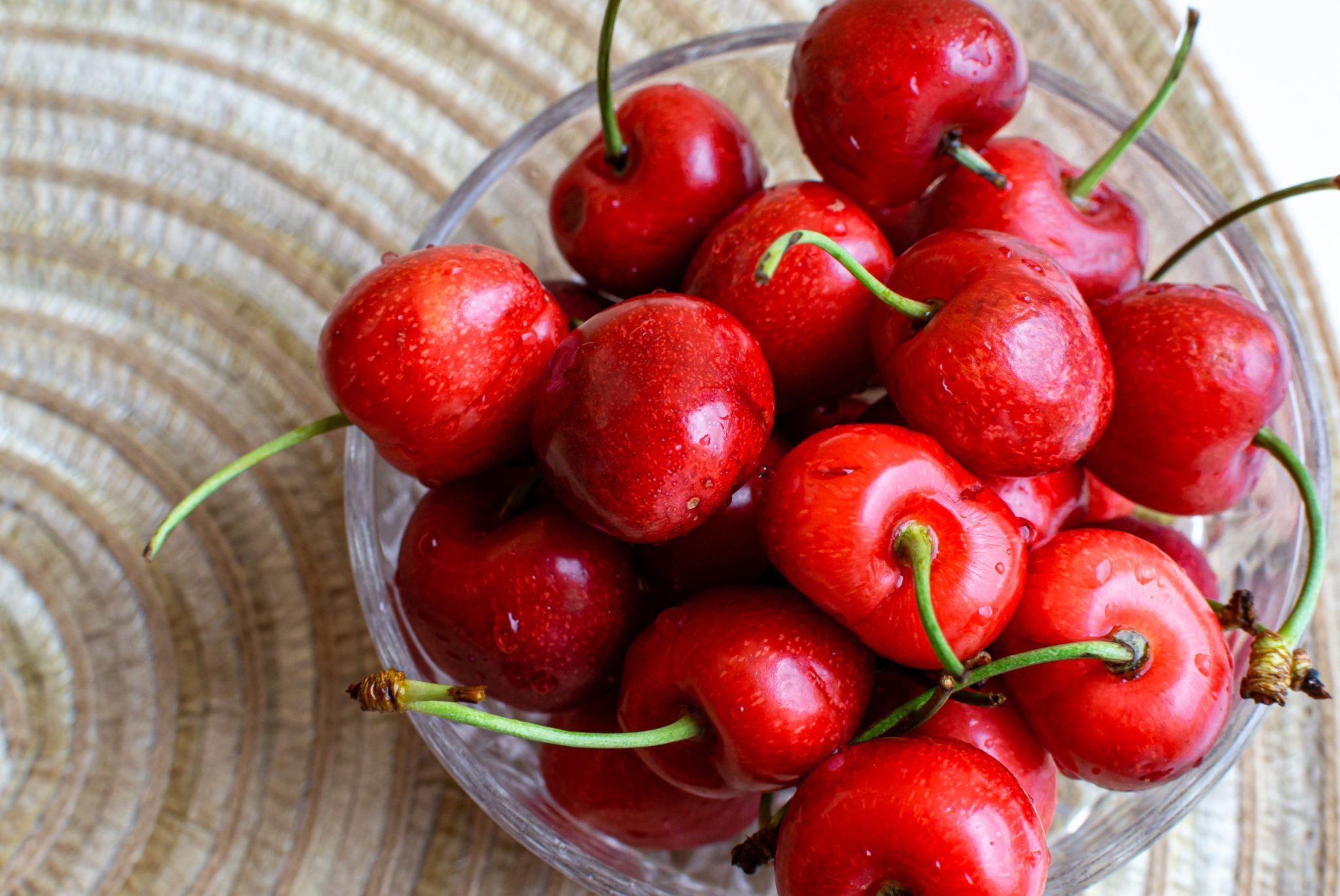 Bright red cherries with green stems in a glass bowl on a wooden surface, close-up view with droplets on fruit.
