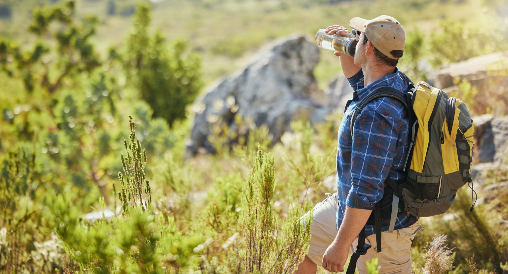 Hiker wearing a blue plaid shirt and tan hat drinks water from a bottle in a sunlit, rocky, green landscape with a yellow backpack on.