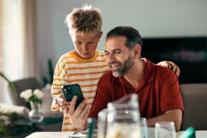 Father and son look at a smartphone together at a dining table in a home setting.