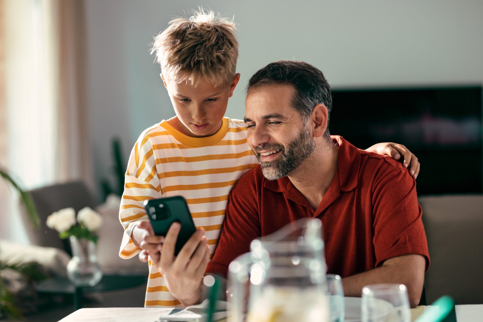 Father and son look at a smartphone together at a dining table in a home setting.