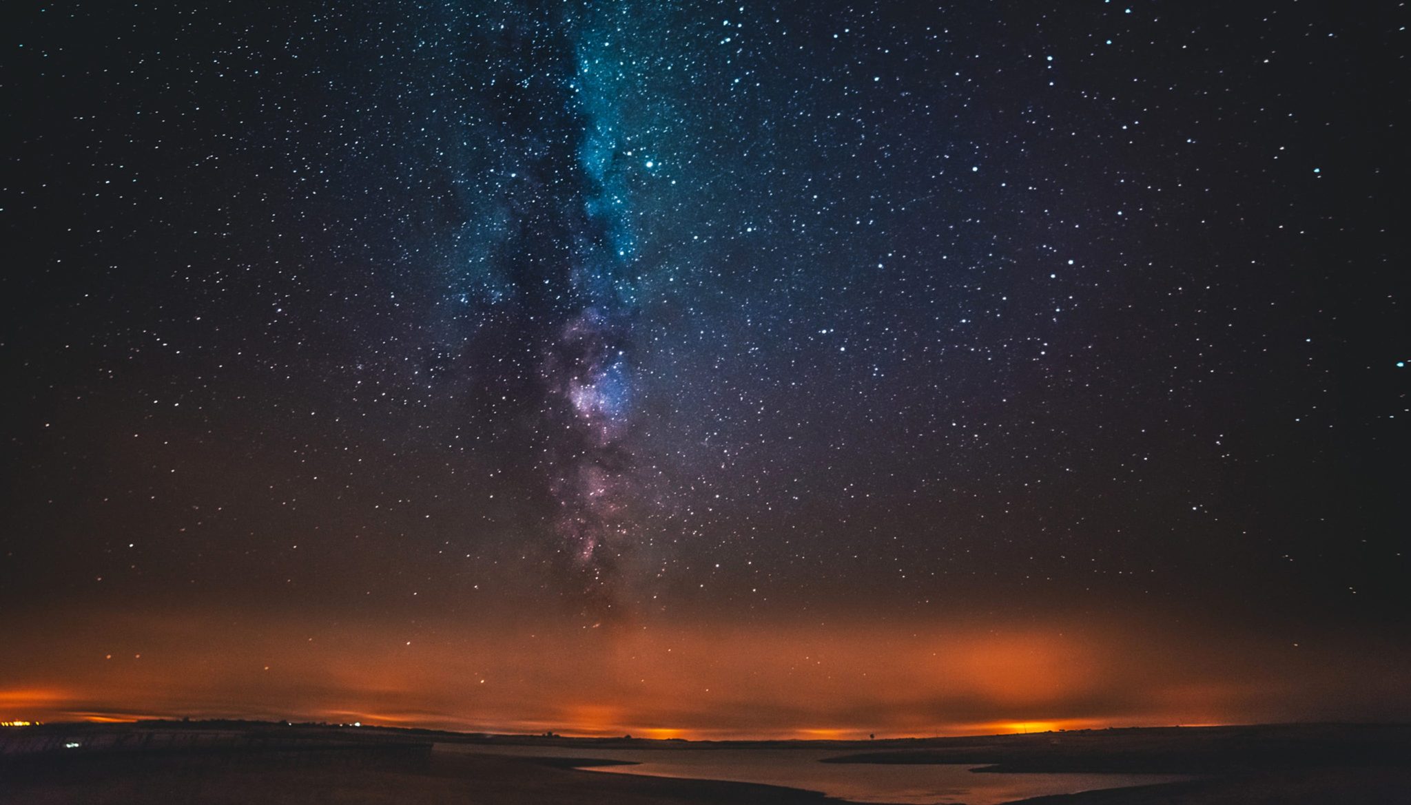 Night sky full of stars including the Milky Way, with a warm orange horizon along a calm coastline at night.
