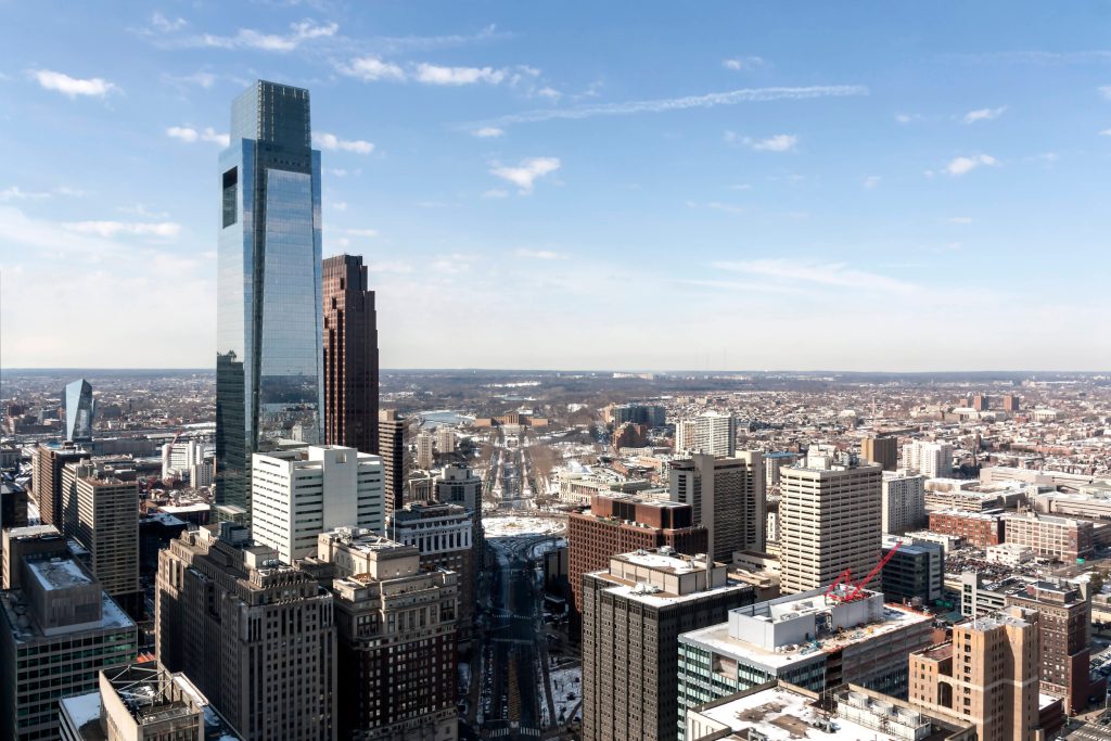 Tall glass skyscraper rises among a dense downtown skyline under a clear blue sky, on a sunny day close to noon.