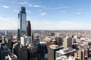 Tall glass skyscraper rises among a dense downtown skyline under a clear blue sky, on a sunny day close to noon.