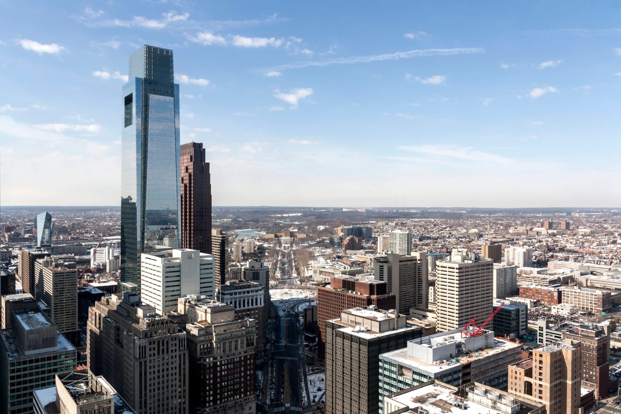 Tall glass skyscraper rises among a dense downtown skyline under a clear blue sky, on a sunny day close to noon.