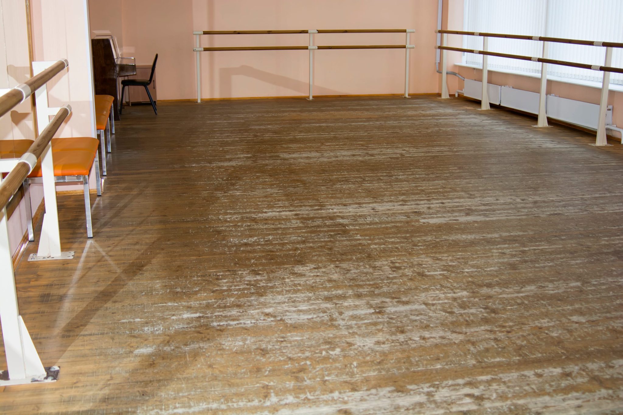 Empty dance studio with worn wooden floor, pink walls, and parallel ballet barres along the walls; chairs and a piano visible in the back corner.