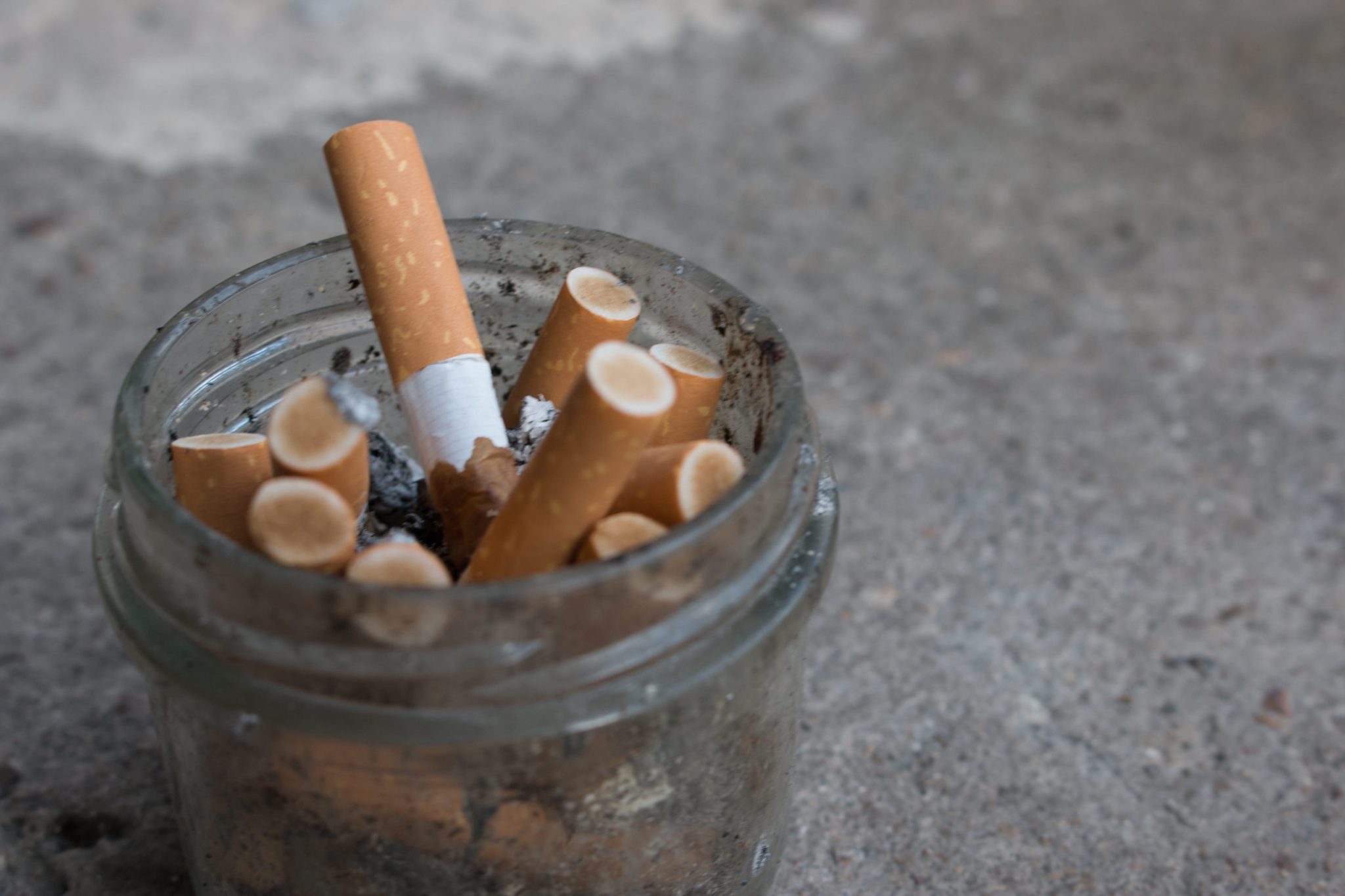 Jar filled with cigarette butts and ash on a rough surface, close-up view.