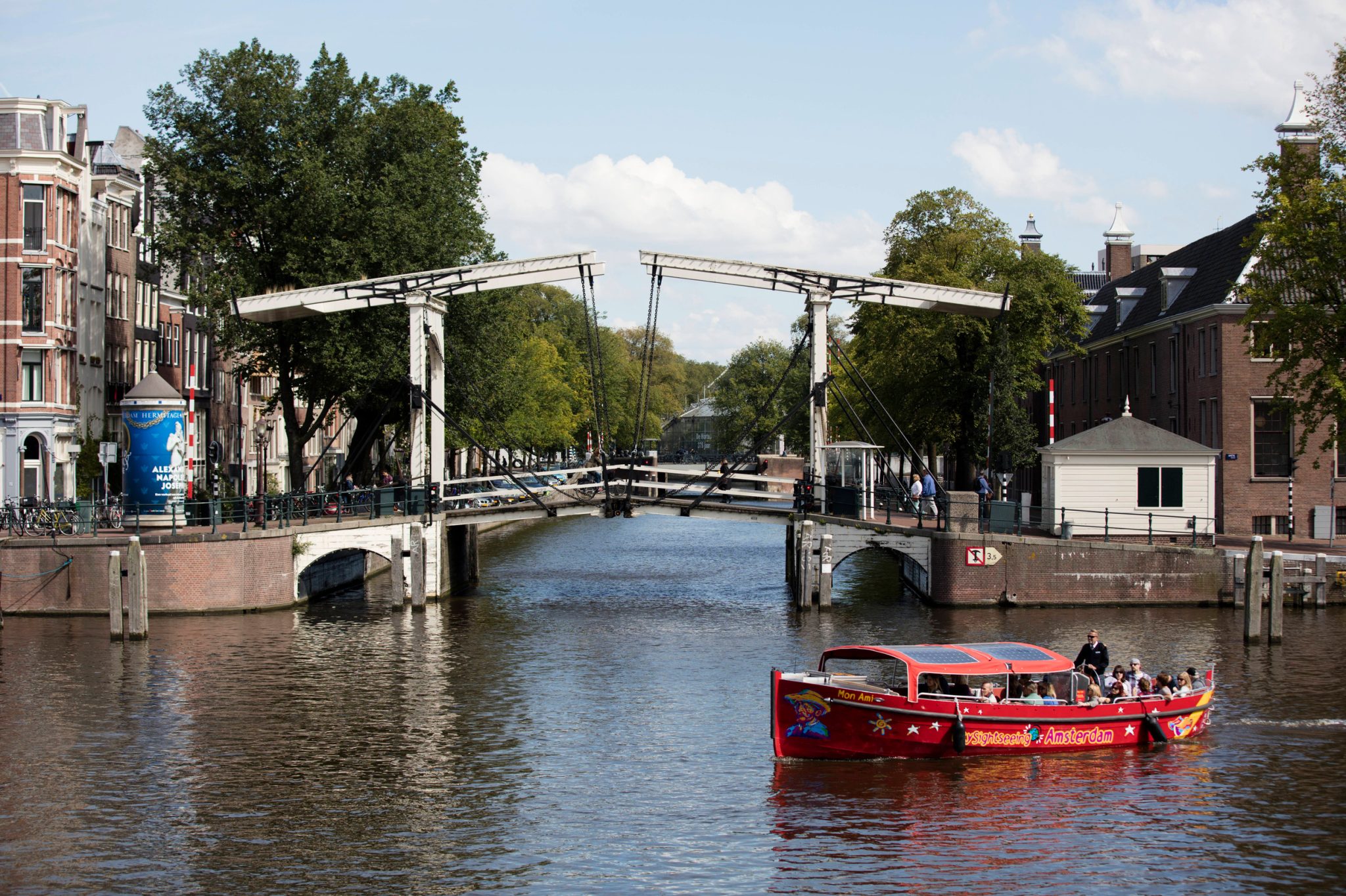 Red sightseeing boat with passengers along a Amsterdam canal, passing under a white drawbridge between brick buildings.