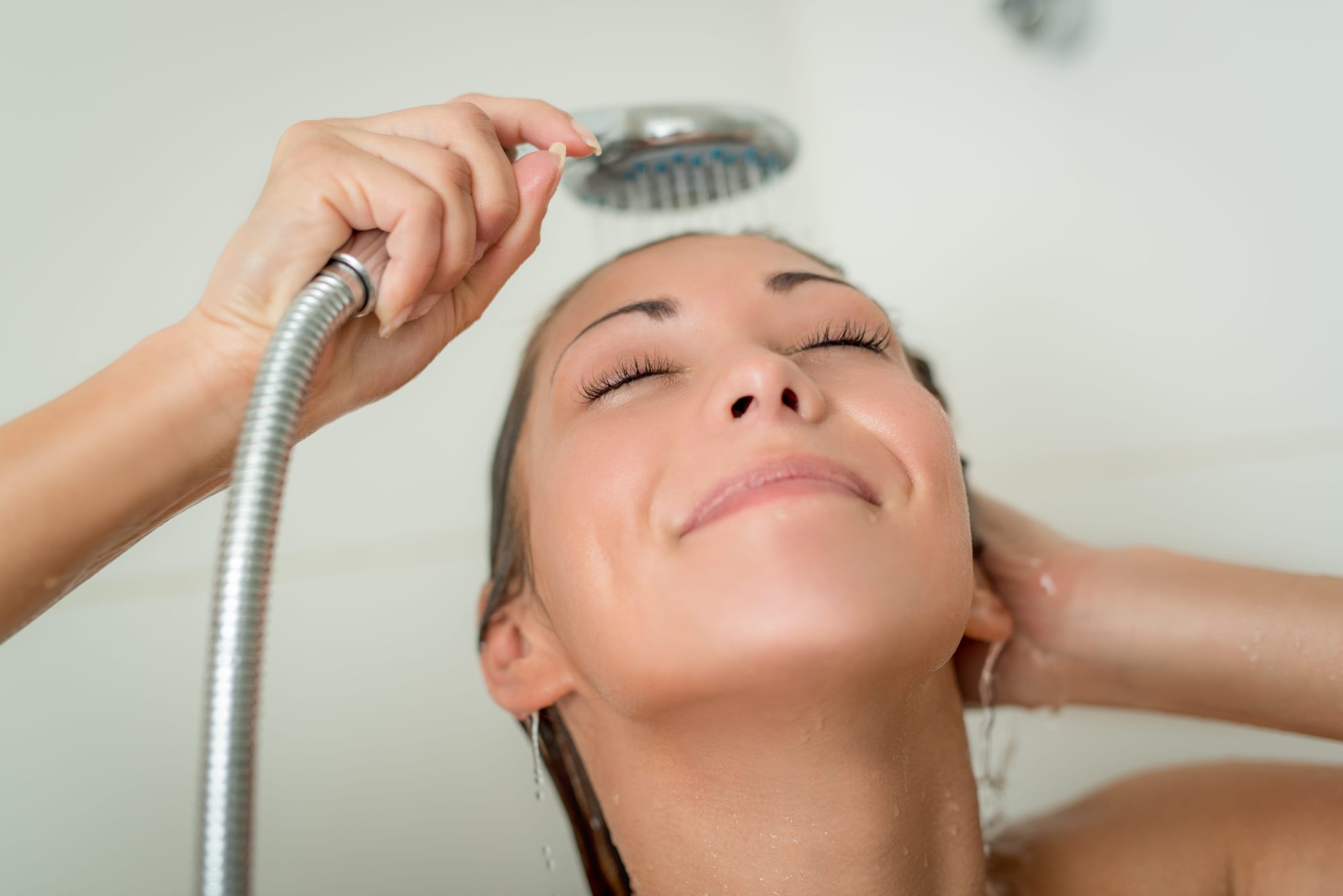 Woman leaning back under a handheld showerhead, rinsing her hair with water.