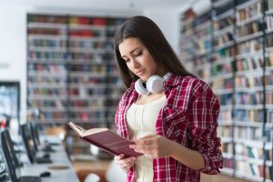 Young woman in a red plaid shirt reads a book in a library, headphones resting around her neck.