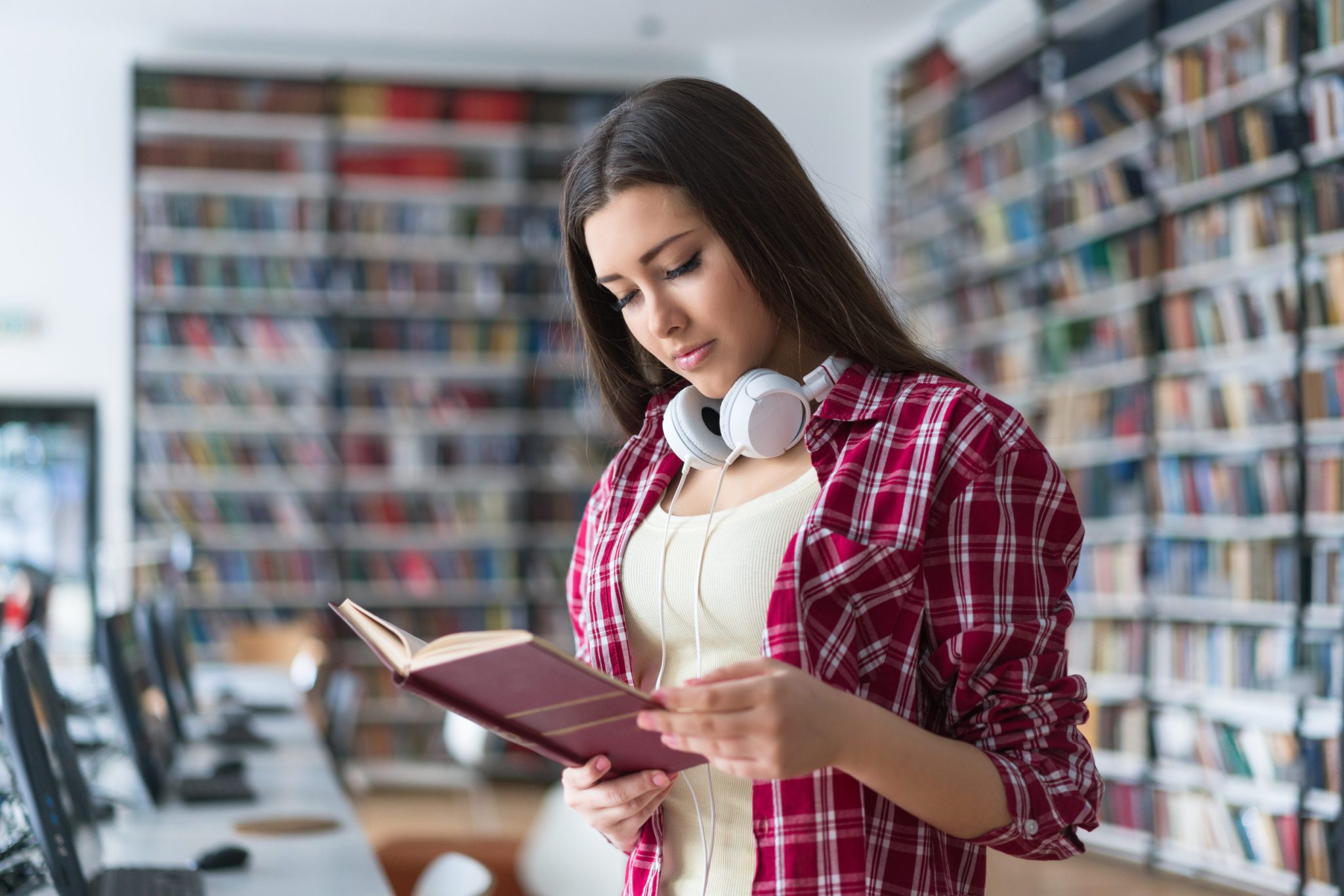 Young woman in a red plaid shirt reads a book in a library, headphones resting around her neck.