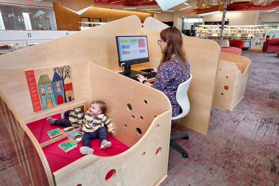Mother works in kid-friendly work carrel next to infant in play area