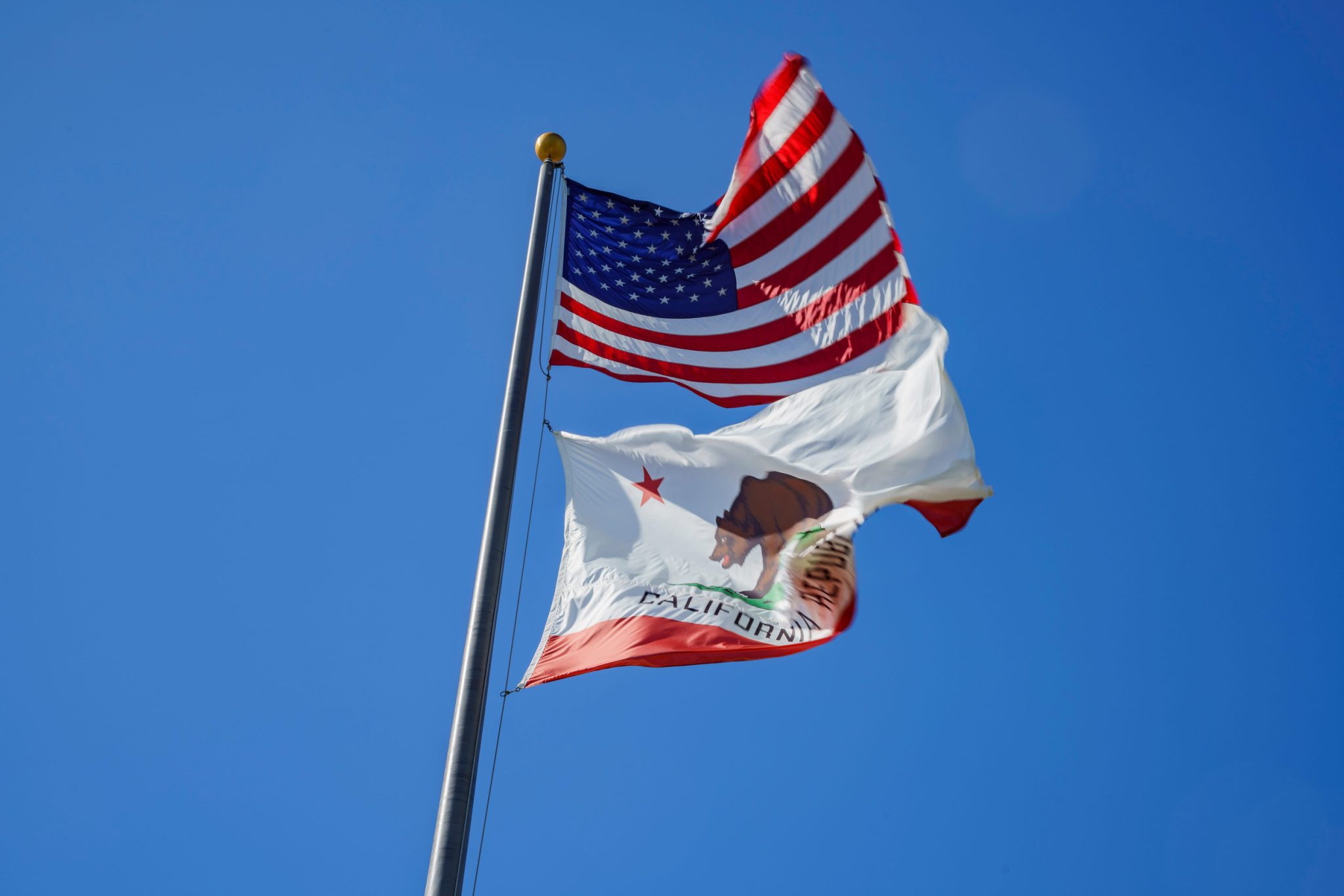 American flag and California state flag waving on a flagpole against a clear blue sky.