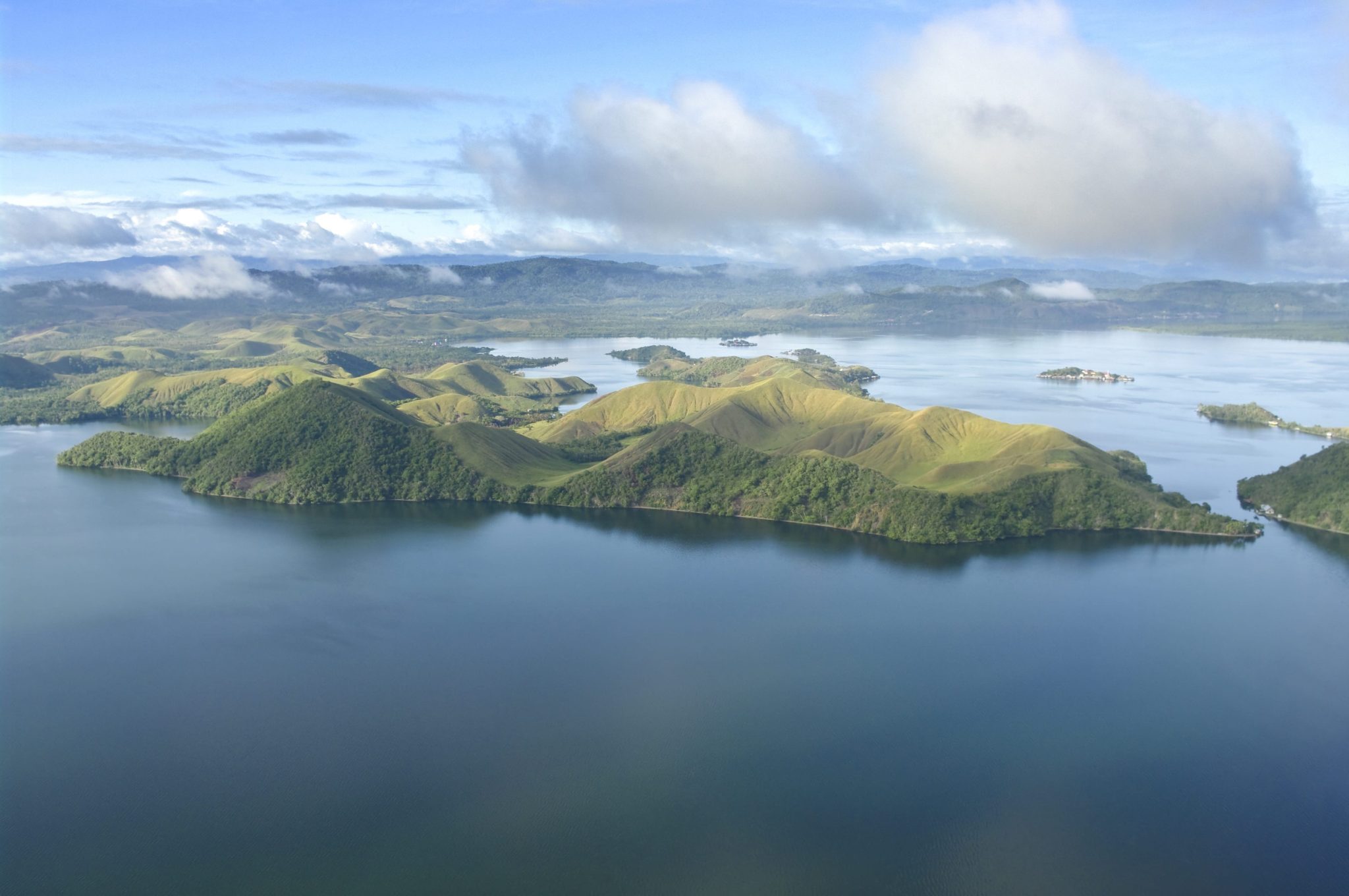 Aerial view of a blue lake with rolling green hills and many small forested islands under a partly cloudy sky, distant mountains on the horizon.