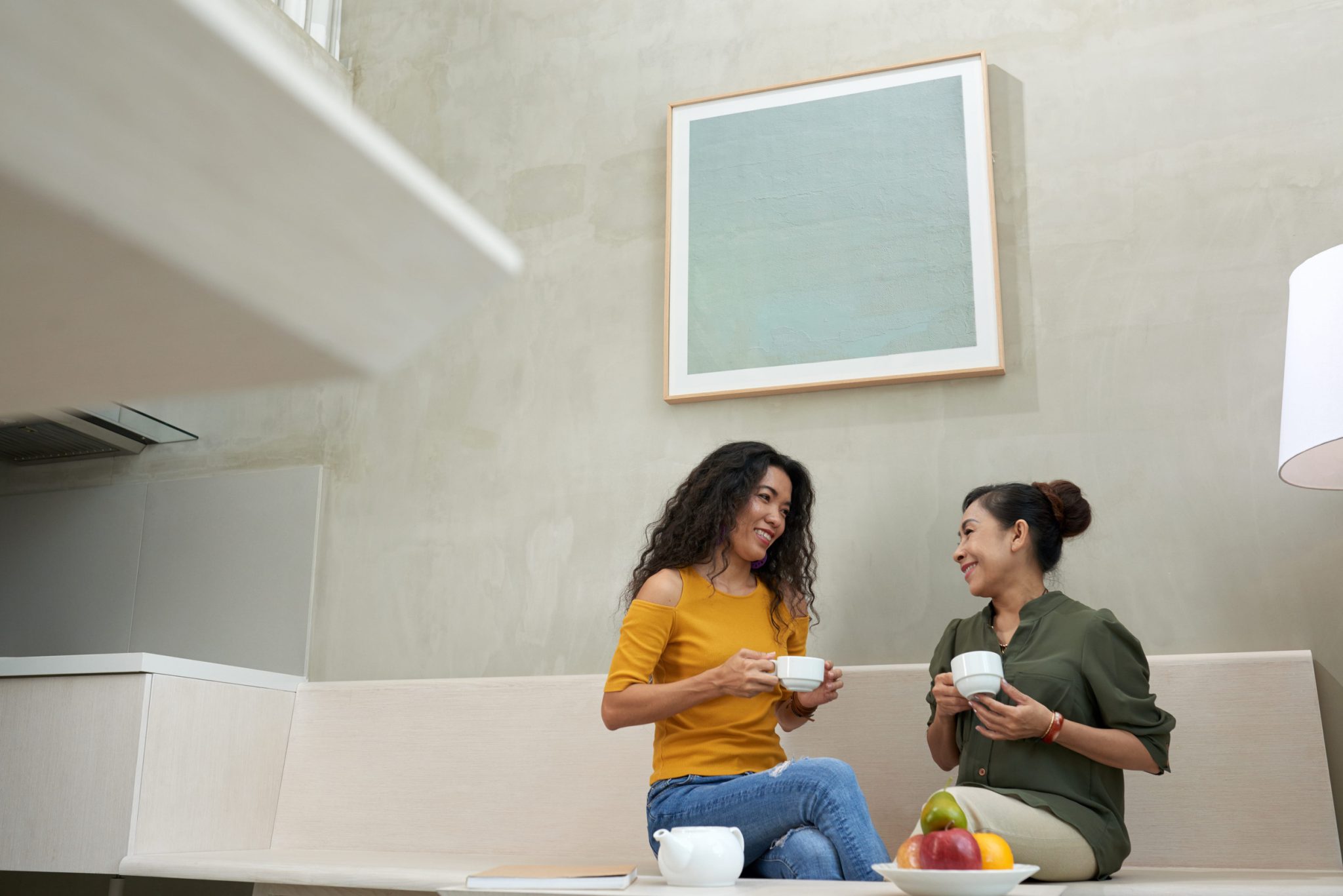 Two women sit on a light wood bench, chatting and holding white coffee cups with fruit on the table in a modern cafe setting.