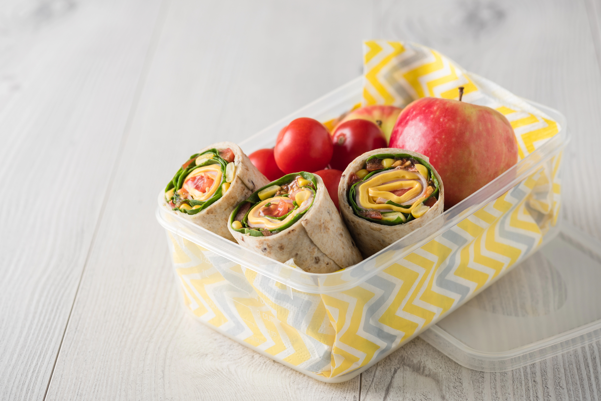 Lunch box with veggie wraps sliced to reveal greens, cheese, and tomatoes, plus cherry tomatoes and a red apple on a yellow chevron napkin.