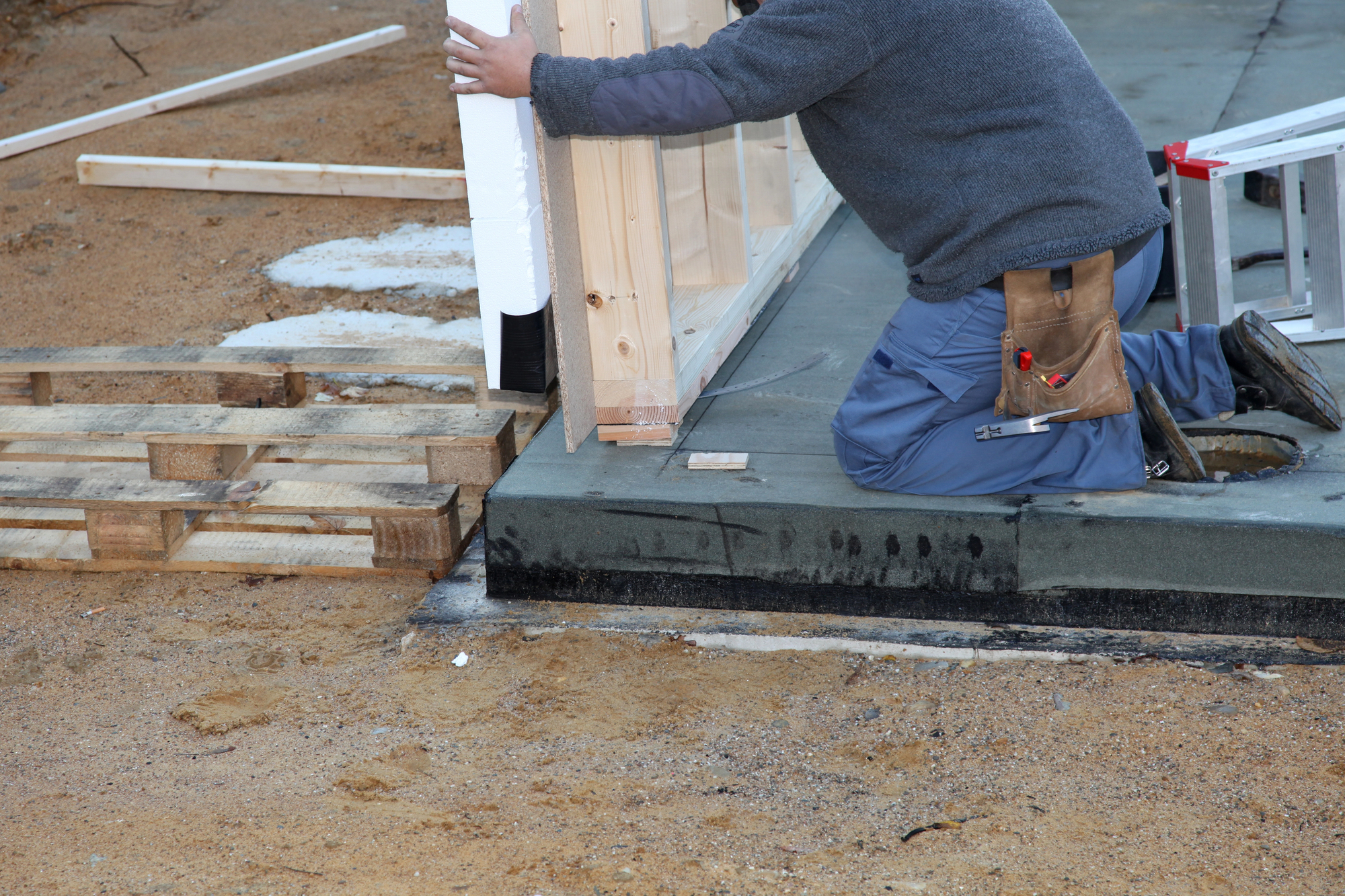 Construction worker kneels on a concrete slab, fastening wooden framing to a wall with tools in a leather belt nearby.