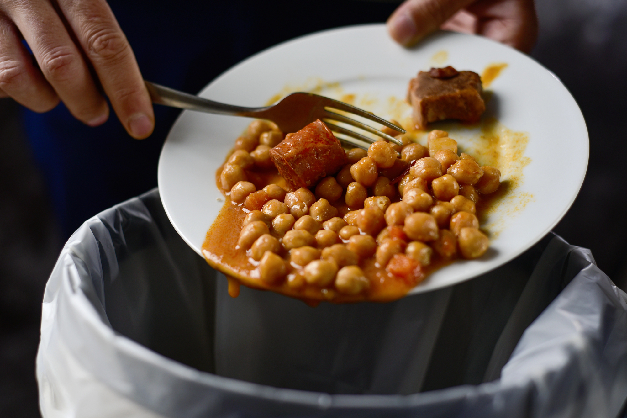 A white plate holds orange tomato-based beans with two chunks of fried meat; a hand grips a fork over the plate, ready to eat.