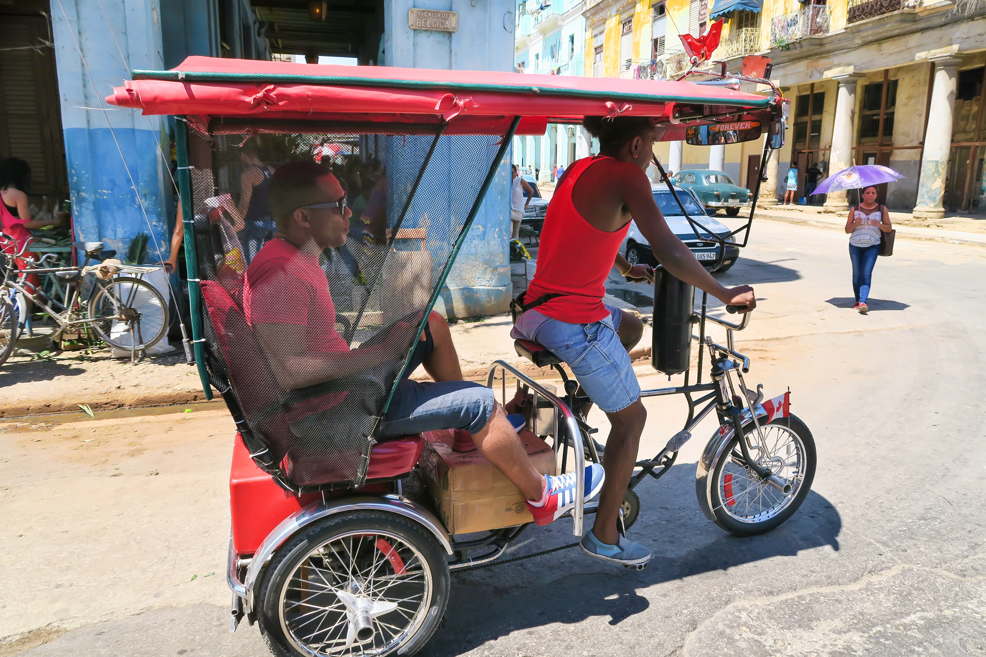 Red tuk-tuk auto rickshaw with mesh passenger seat, two riders cruising down a sunny urban street.