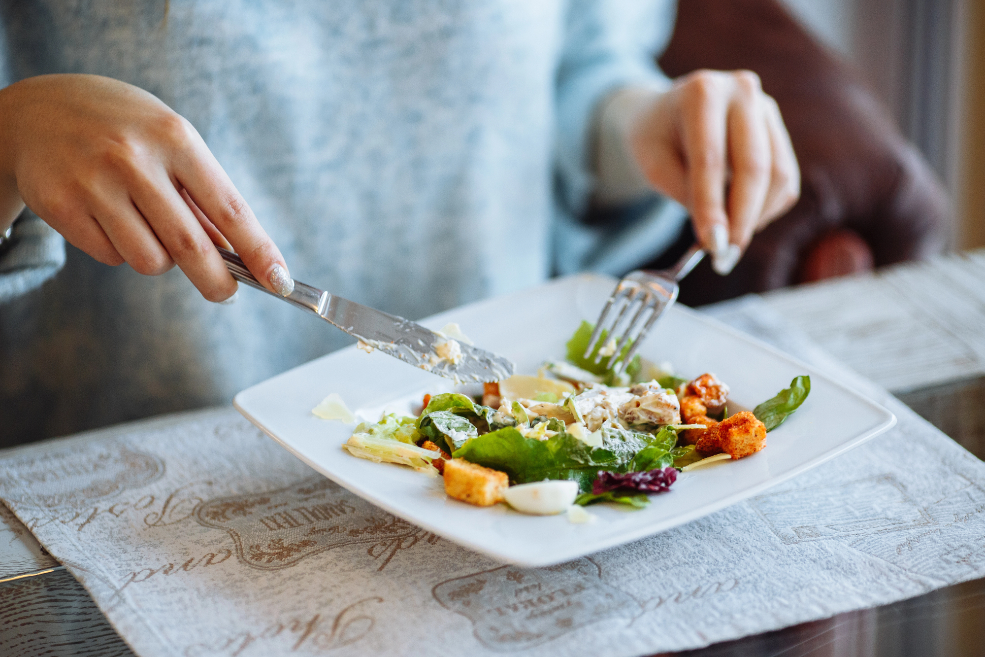Hands cutting a mixed salad with a knife and fork on a square white plate.