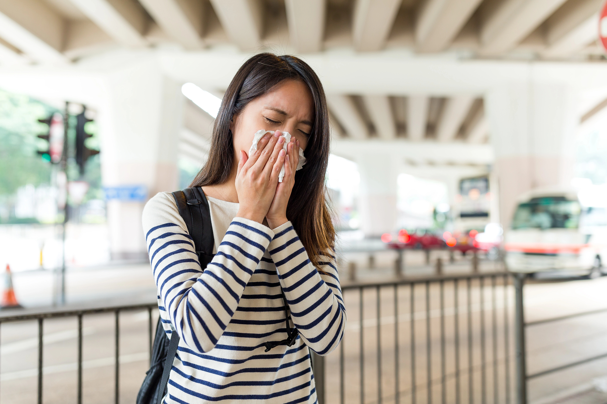Woman in a striped shirt sneezing into a tissue in an outdoor urban area under a highway overpass.