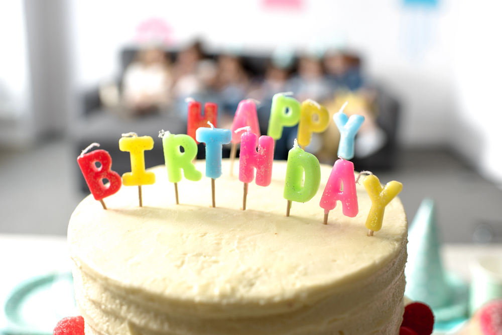 Birthday cake topped with multicolored letter candles spelling 'BIRTHDAY'; party guests blurred in the background.