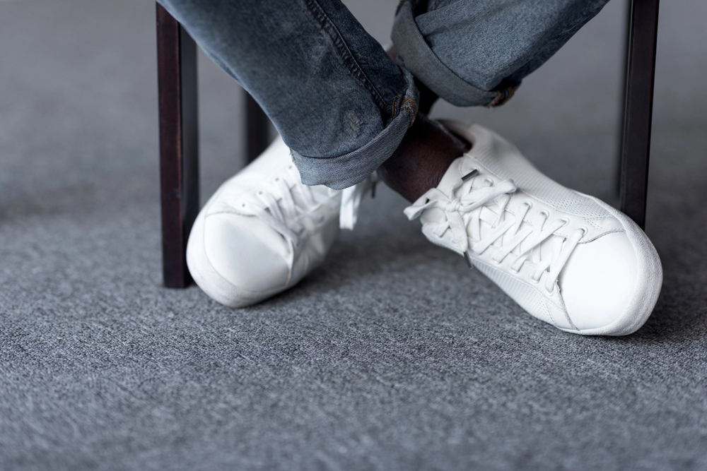 Person seated with crossed legs, wearing white sneakers and rolled cuffs jeans on a gray carpeted floor under a chair leg.