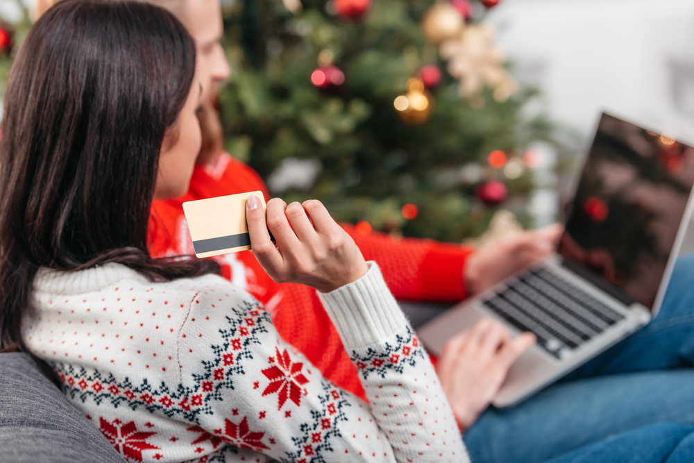 Couple relaxing at home by a Christmas tree; woman holds a gold card with a dark stripe while a man uses a laptop nearby.