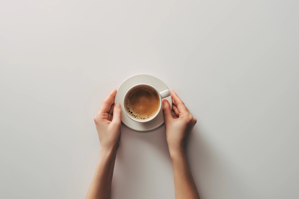 Top-down view of two hands holding a white cup of coffee on a saucer against a pale background, crema visible on the surface.