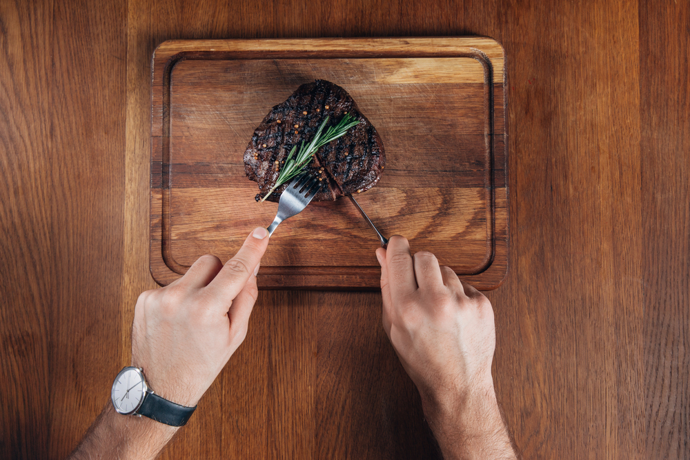 A person carves a grilled steak with a fork and knife on a wooden cutting board, rosemary garnish on top of the meat.