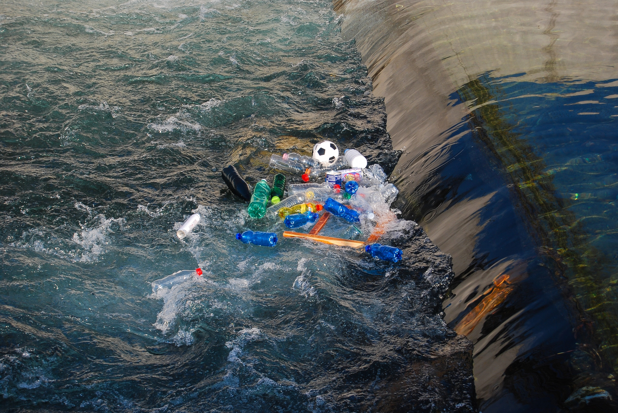 Plastic pollution in a rocky shoreline pool with empty bottles, cans, and a soccer ball among the debris.