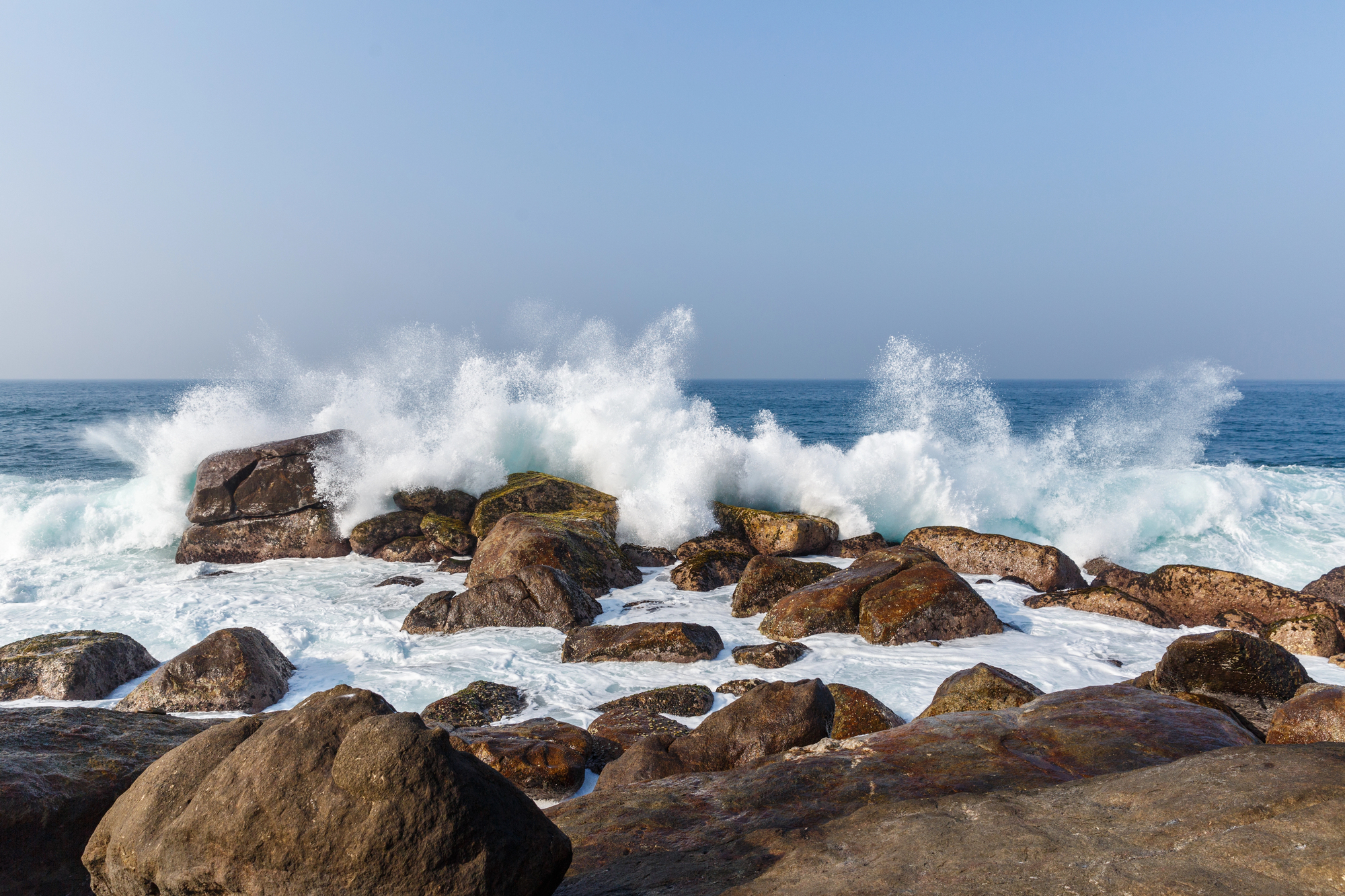 Rugged rocky shore with crashing waves spraying white foam under a clear blue sky, ocean in the background