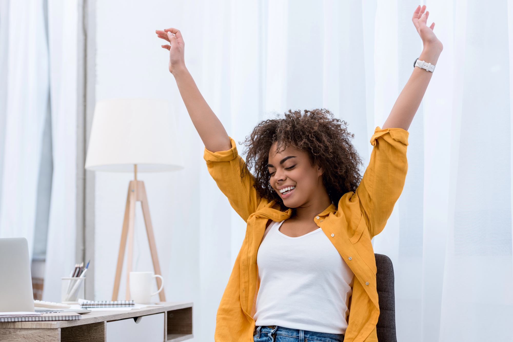 Woman with curly hair in a mustard shirt raising arms in celebration in a bright home office setting with a laptop nearby.