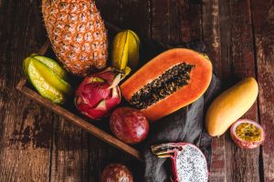 Assorted tropical fruits on a dark wooden surface: pineapple, papaya half, dragon fruit, mango, passion fruit, and a red fruit (pomegranate) in a rustic display.