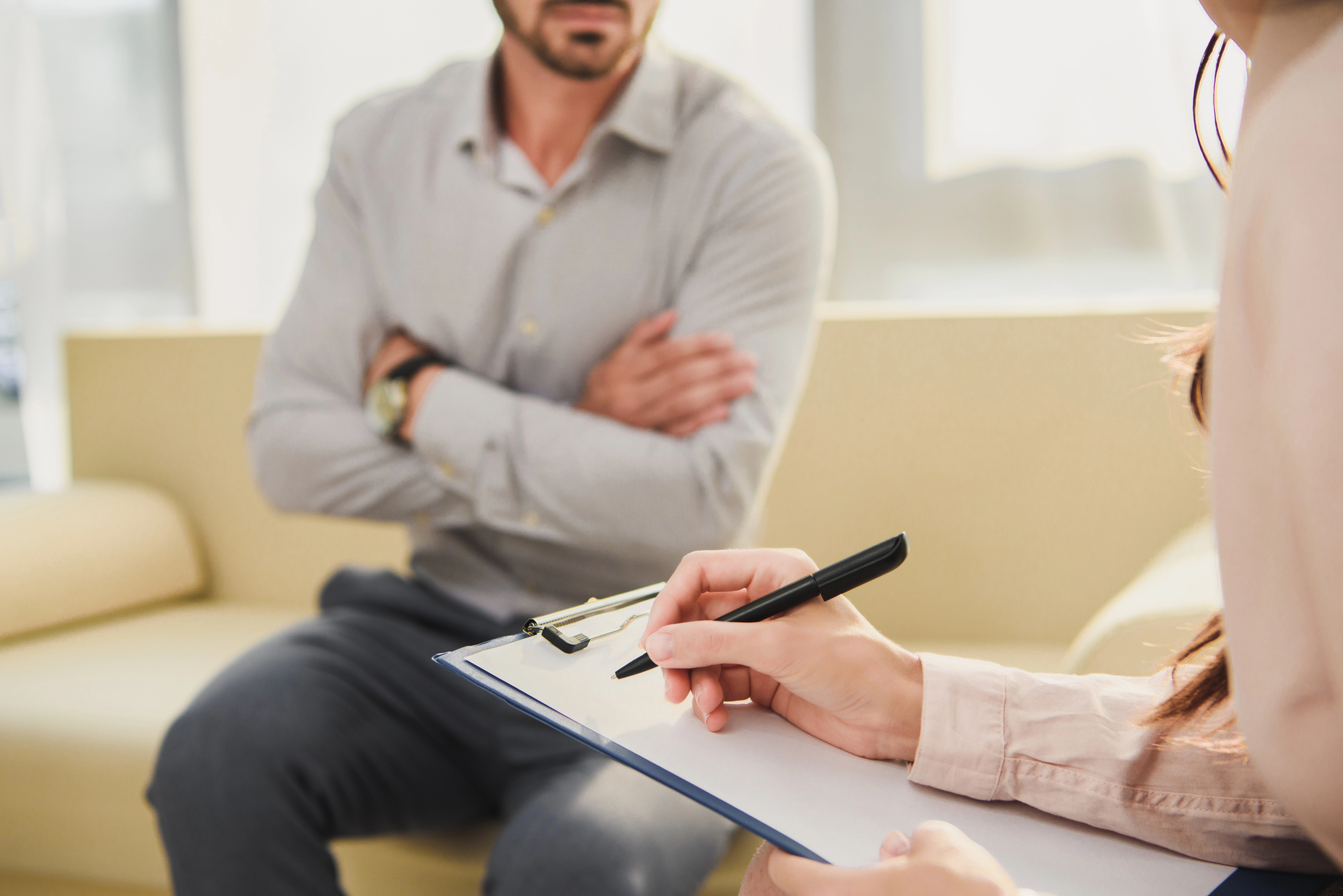 Close-up of hands writing on a clipboard during a consultation, with a seated person blurred in the background in a bright waiting area.