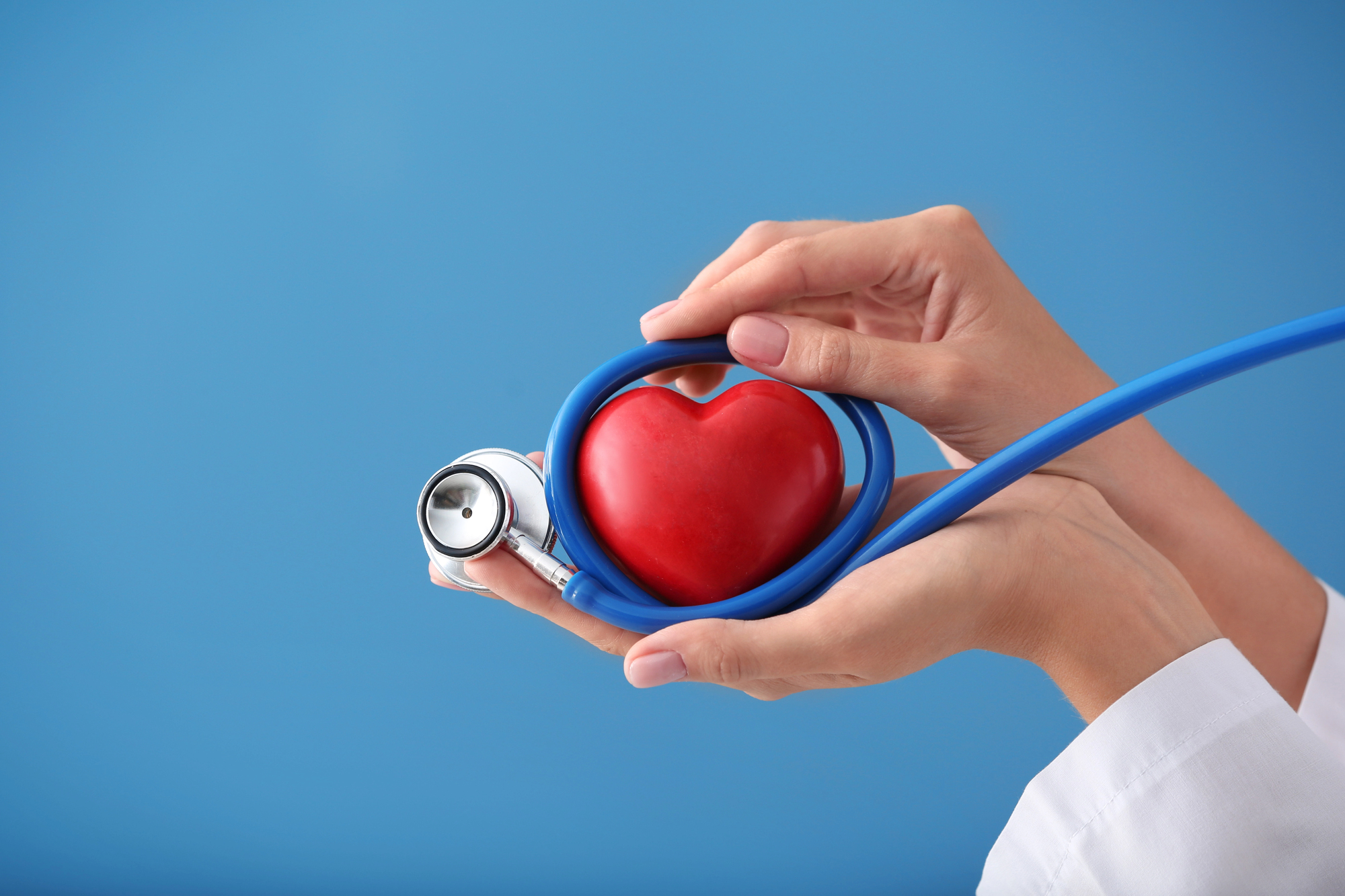 Hands hold a red heart with a blue stethoscope, blue background, symbolizing heart health and medical care,
