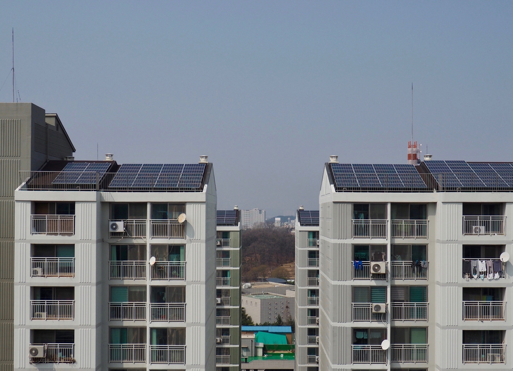 Row of white apartment buildings with numerous solar panels on flat roofs, balconies, and satellite dishes against a clear blue sky.