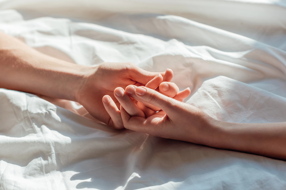Two hands gently holding each other on a white bedsheet, conveying comfort and connection.
