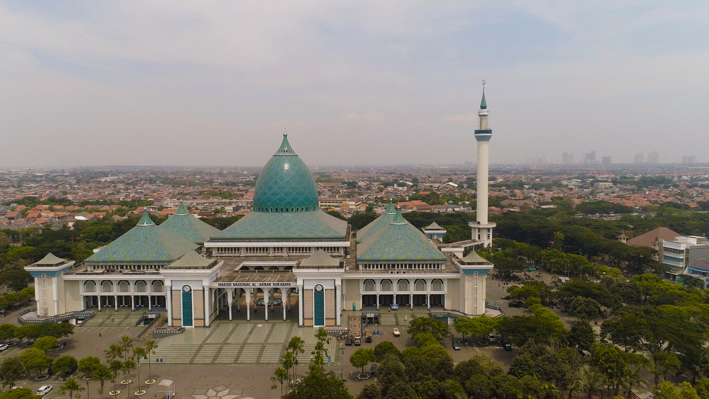 Surabaya's National Mosque Al-Akbar with turquoise dome and tall minaret, surrounded by trees and city skyline.