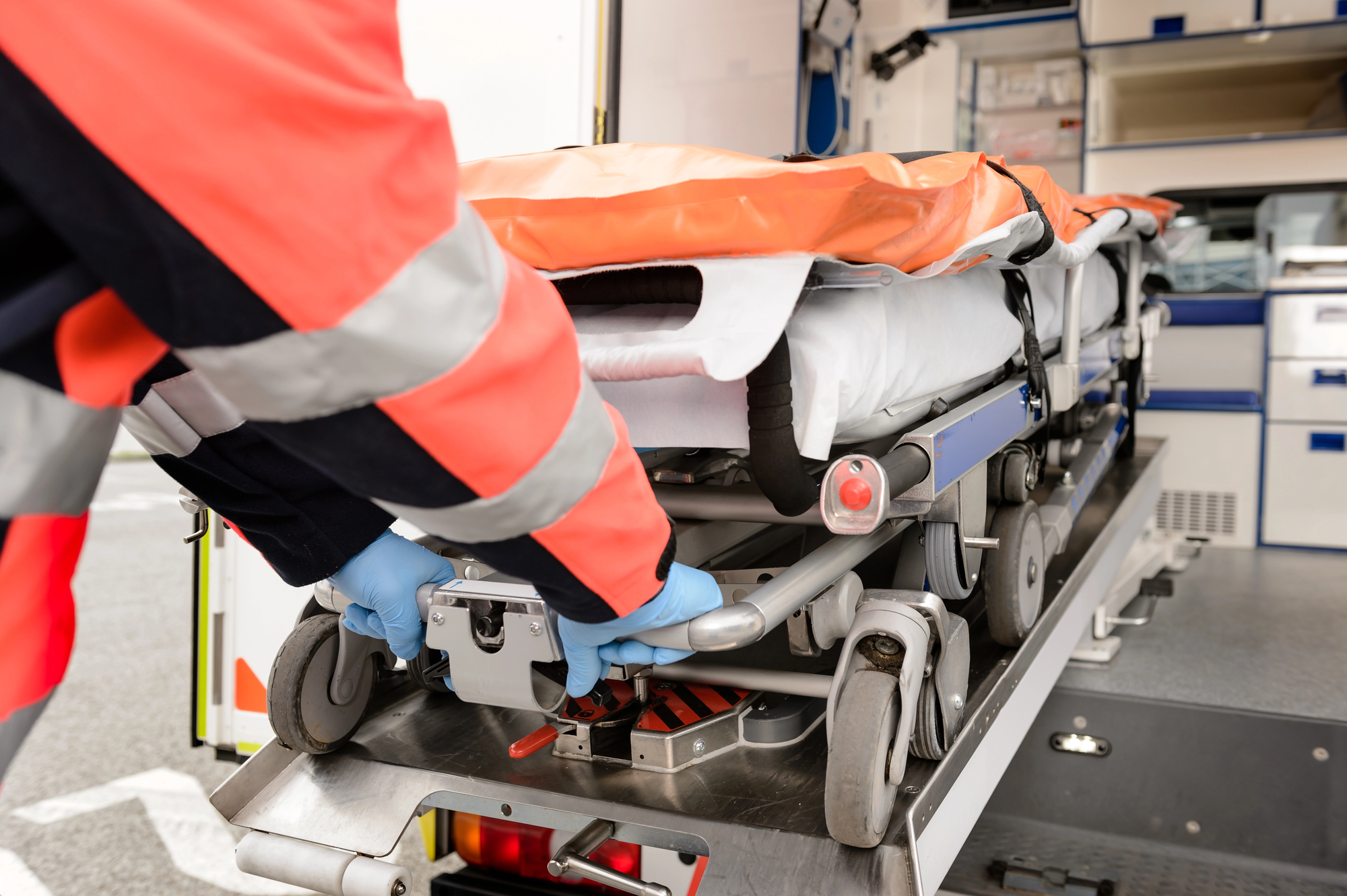 Emergency medical technicians loading a wheeled stretcher into an ambulance, wearing high-visibility orange jackets and blue gloves.