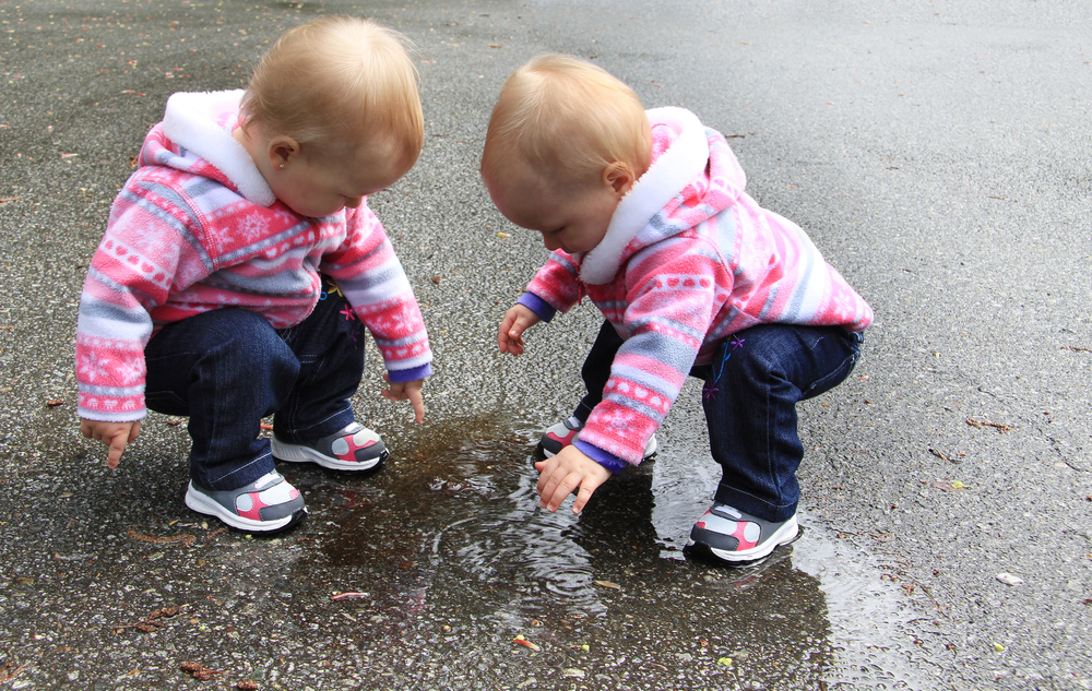 Two toddlers in pink striped hoodies crouch by a puddle on wet pavement, touching the water with their fingers.